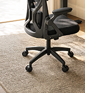 Black ergonomic office chair with mesh back and padded seat, shown on light carpet with wooden flooring and furniture visible in background.