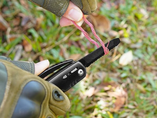 Close-up of hands using pliers to attach a pink cord to a black tactical tool or device in an outdoor setting.