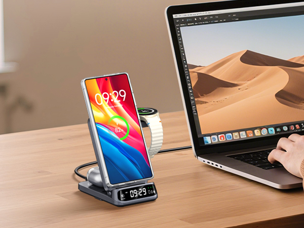 Smartphone charging stand on desk, displaying colorful screen with time. Features built-in digital clock. Positioned next to laptop showing desert landscape image.