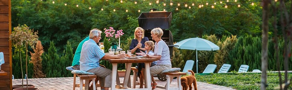 Outdoor patio dining scene with round wooden table under string lights, featuring garden furniture and blue lounge chairs in background.