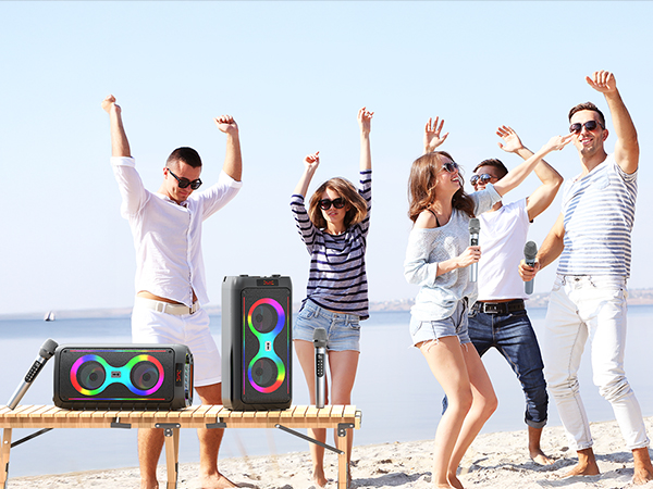 Group of people dancing on beach with portable speakers, wearing casual summer attire against ocean backdrop.