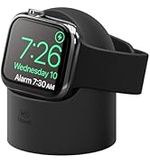 Smartwatch with square display showing time 7:26 and date Wednesday 10, resting on a black charging stand. Watch has black band and silver frame.