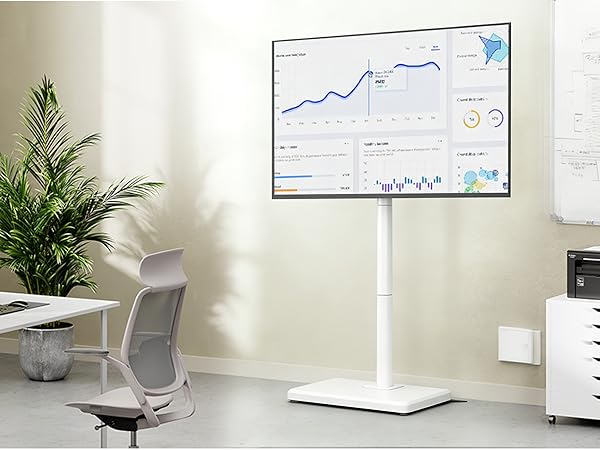 Modern office workspace with white adjustable desk, grey ergonomic chair, wall-mounted analytics displays, and decorative plants. Clean minimalist design with natural lighting.