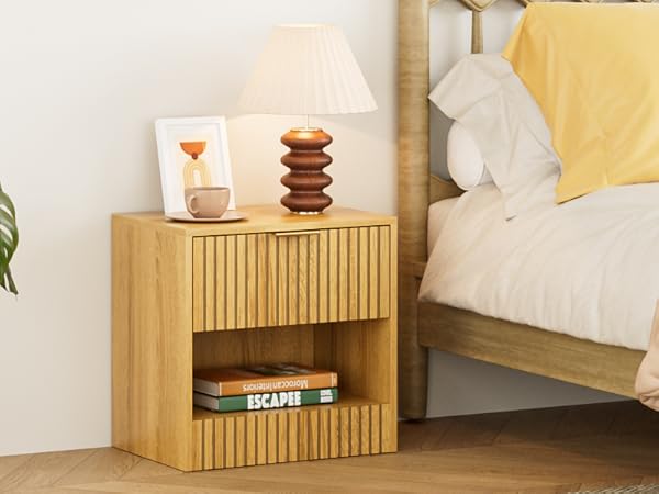 Wooden nightstand with ridged front panel and open shelf, placed beside a bed with yellow pillows. Features table lamp and small decor on top.