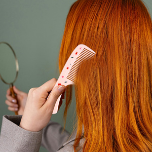 Person holding white hair styling tool against long, vibrant orange-red hair. Tool appears to be a hair straightener or styling device with white cord visible against neutral background.