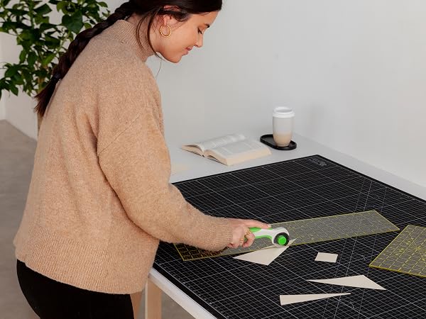 Person working at a black textured cutting mat on white surface, using measuring tools with green accents. Houseplant and glass of water visible in background.
