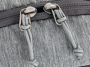 Close-up of two metal zipper pulls on a gray textured fabric, showing the zipper mechanism and pull tabs.