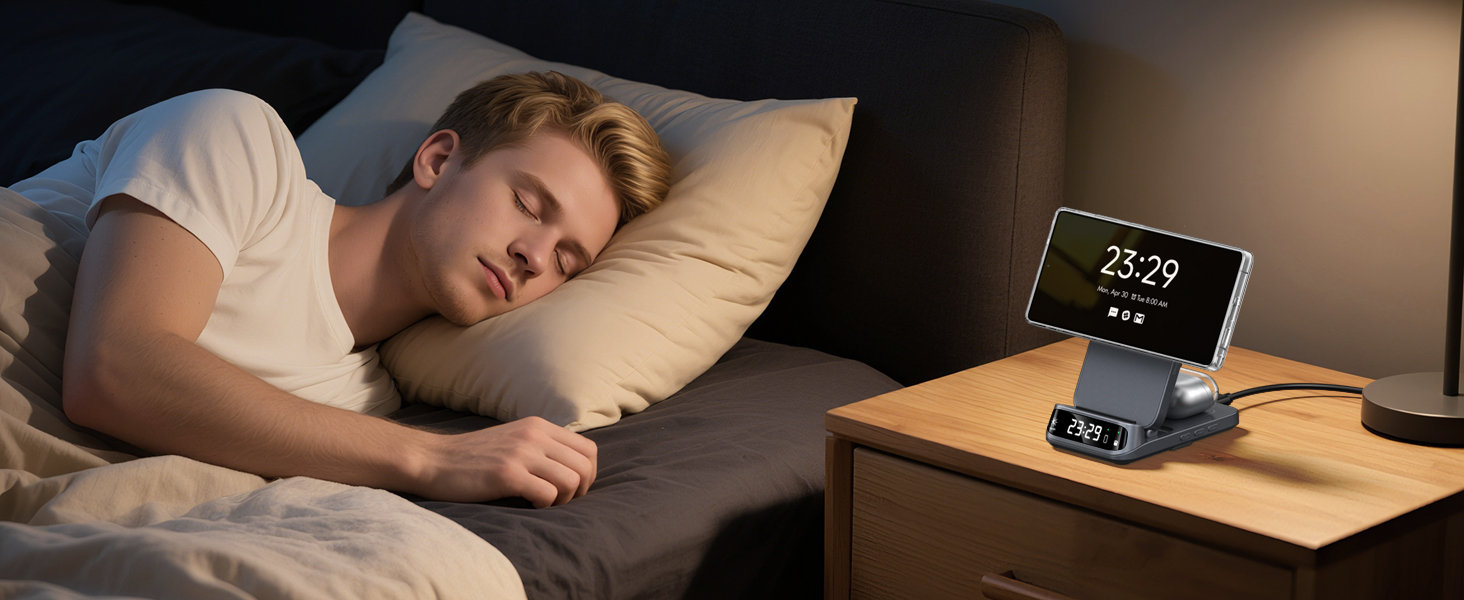 Electronic device on nightstand displaying time 23:29. Device has a stand for holding a smartphone. Person sleeping in bed nearby.