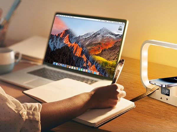 Desk setup with laptop, curved LED lamp, and wireless charging pad. Person's hands visible, working in a home office environment.