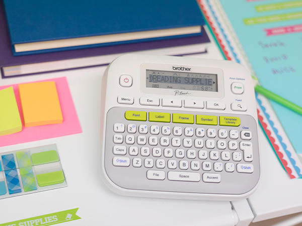 Brother P-touch PTD210SV Label Maker on desk with notebooks, papers. Screen reads READING SUPPLIES