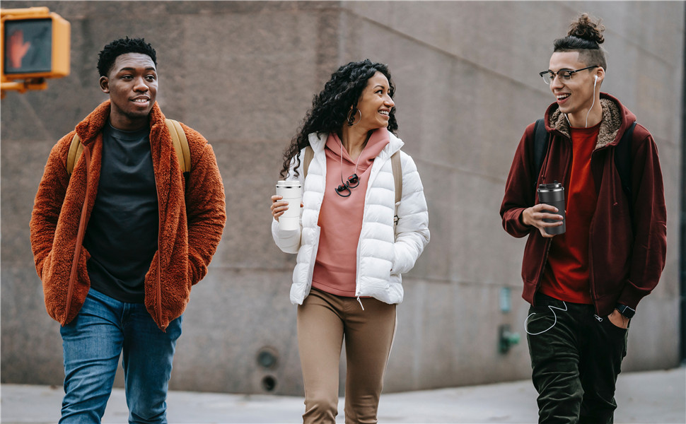Three young adults walking outdoors, wearing winter jackets. One in orange fleece, one in white puffer, and one in maroon jacket. Urban setting visible.