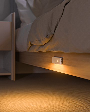 Small motion-activated night light mounted under a bed frame, casting warm illumination onto carpet below.