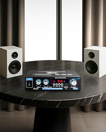 Home audio system on black table: compact amplifier with digital display, volume controls, and input options, flanked by two white bookshelf speakers with visible drivers.