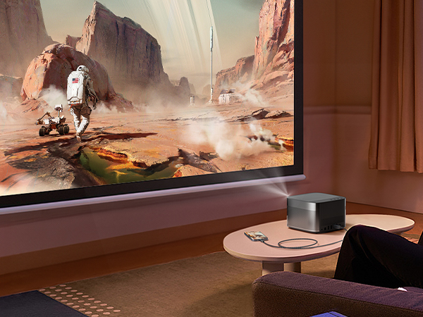Home theater setup showing a compact black projector on wooden table displaying a sci-fi desert scene, with ambient room lighting.