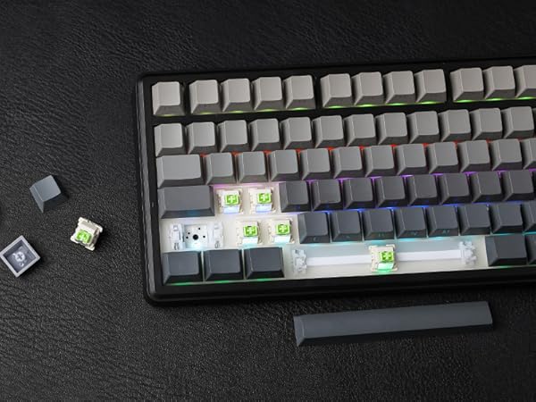 Mechanical keyboard with white keycaps and RGB lighting. Several keycaps removed, revealing green switch mechanisms. Detached spacebar and loose keycaps visible on dark surface.