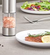 Stainless steel salt or pepper grinder with pink Himalayan salt visible through transparent chamber, shown on table with dining setting in background.