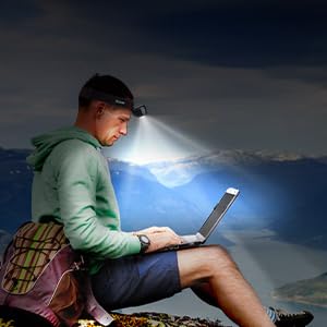 Someone using a laptop outdoors at night on a rocky surface, illuminated by a bright headlamp against a dark mountain landscape backdrop.