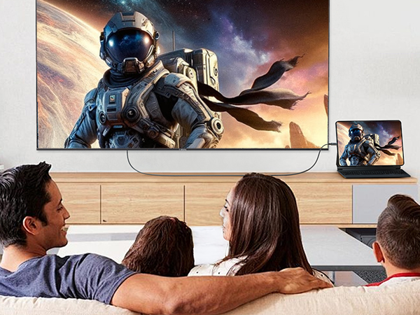 Large flat-screen TV displaying space-themed game artwork, connected to laptop. Four people seated on couch, viewing the screen together.