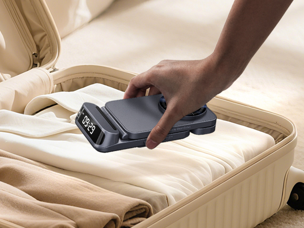 Hand placing a small, rectangular digital luggage scale on folded clothes inside an open suitcase. Scale displays '0.00' on its screen.