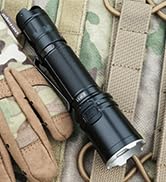 Black tactical flashlight mounted on camouflage material, showing robust metal construction and mounting clips.
