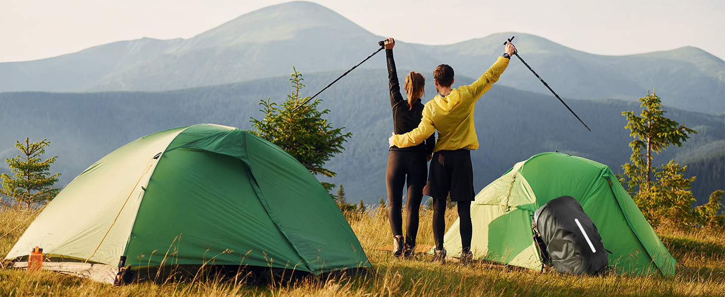 Two green camping tents set up on a mountain ridge, with people in yellow jackets standing between them against a backdrop of misty mountains.
