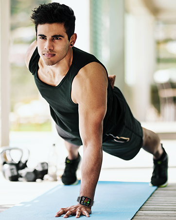 Man in black tank top and shorts performing push-up exercise on blue yoga mat. Indoor setting with natural lighting.