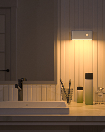 Modern bathroom vanity with motion-sensor LED light mounted above white paneled wall, featuring sink and green glass containers.