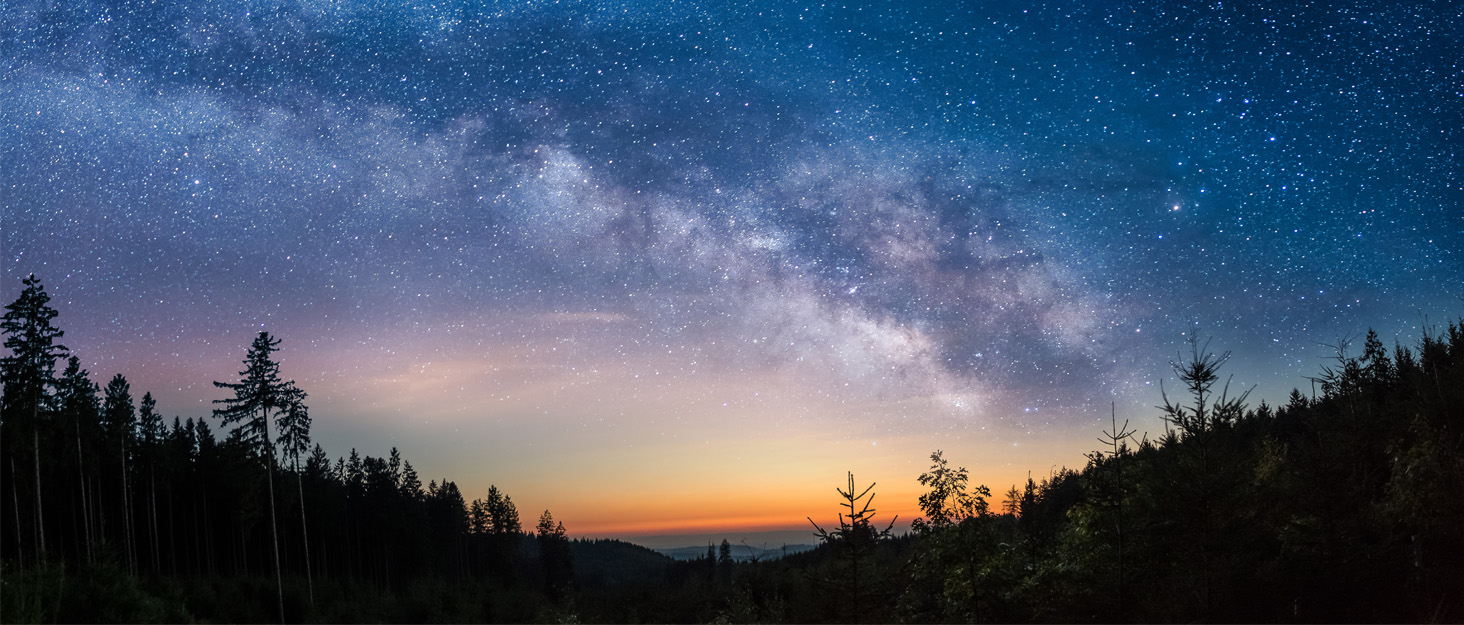 Night sky landscape with Milky Way visible over silhouetted forest. Horizon shows faint glow of sunset or sunrise.