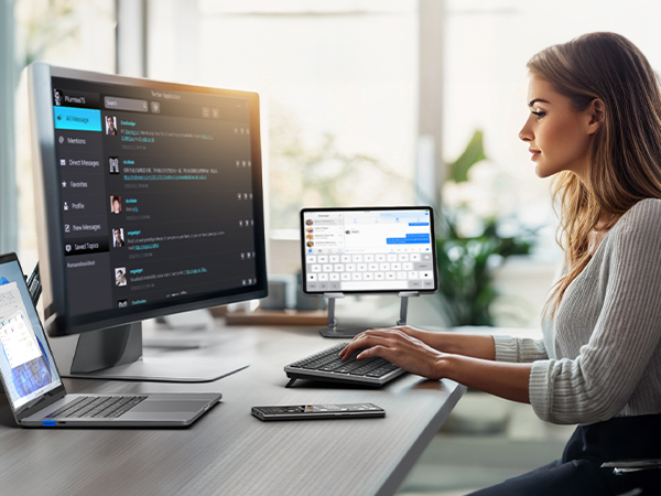 Professional workspace setup with multiple monitors displaying communication software interface, laptop, and keyboard on a wooden desk.