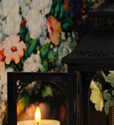 Dark atmospheric scene with candlelight illuminating floral elements in the background.