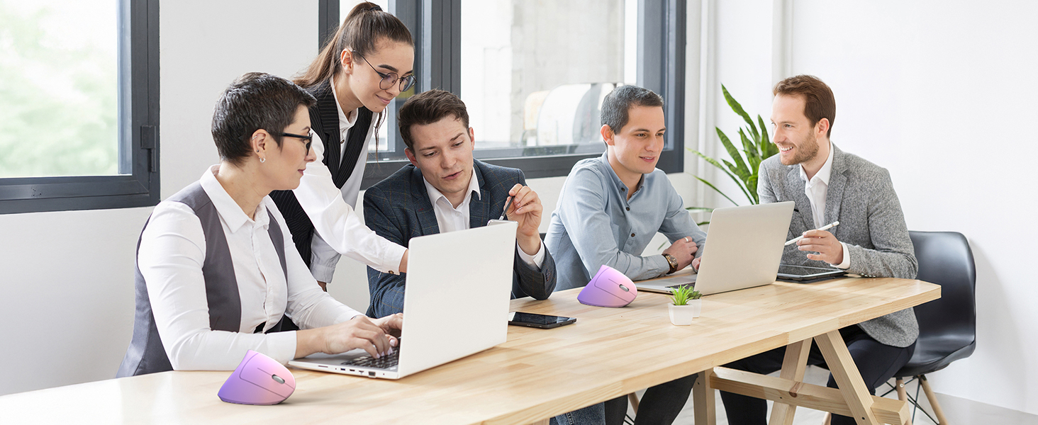 Professional workspace showing business team collaborating at long wooden desk with laptops and decorative pink spherical objects, in bright modern office with window.