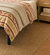 Bed corner with patterned tan and cream striped bedding and pillows on wooden flooring.