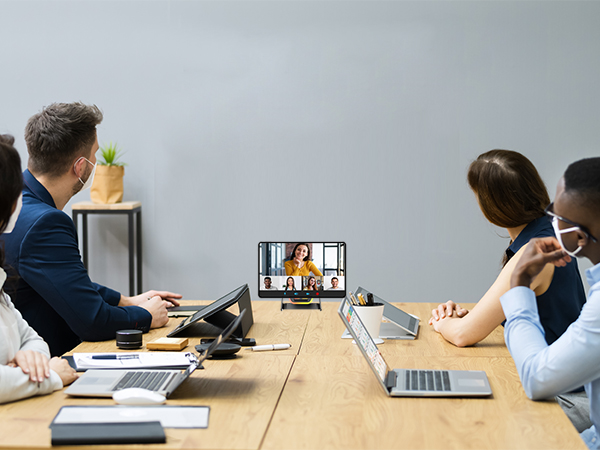 Business meeting scene showing people seated at conference table with laptops and tablets, viewing content on screen during video call.