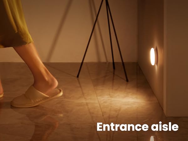 Illuminated motion sensor light on wall near floor, activating as person in slippers walks by. Warm glow on wooden floor, labeled 'Entrance aisle'.