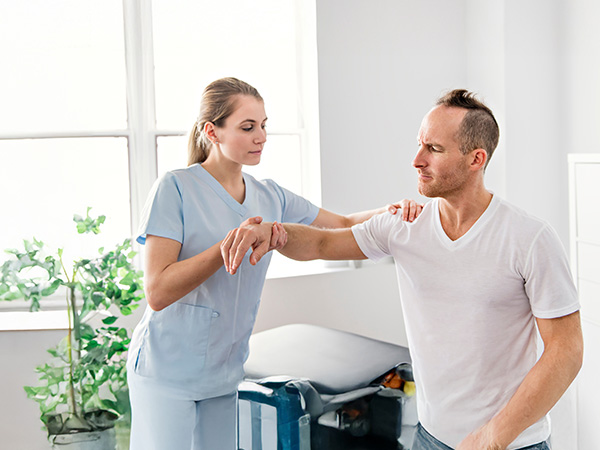 Healthcare provider in light blue scrubs examining patient's arm/shoulder mobility in clinical setting with natural lighting and indoor plants visible.