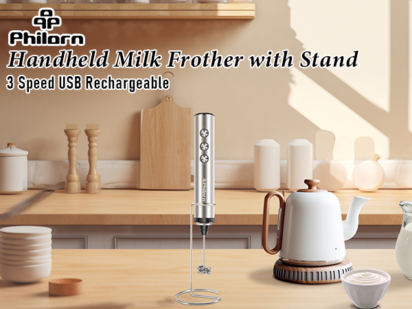 Stainless steel handheld milk frother with stand on wooden surface. White ceramic cups and kettle visible. USB rechargeable with 3 speeds.