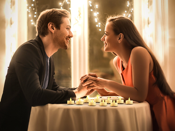 Romantic table setting with multiple lit tea light candles. Two people lean towards each other across the table in a warm, intimate atmosphere with string lights in the background.