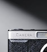 Text reads 'CAMERA'. Close-up detail of what appears to be a camera or electronic device with textured black surface and metallic elements.
