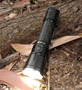 Black tactical flashlight illuminated and positioned on ground with fallen leaves visible.