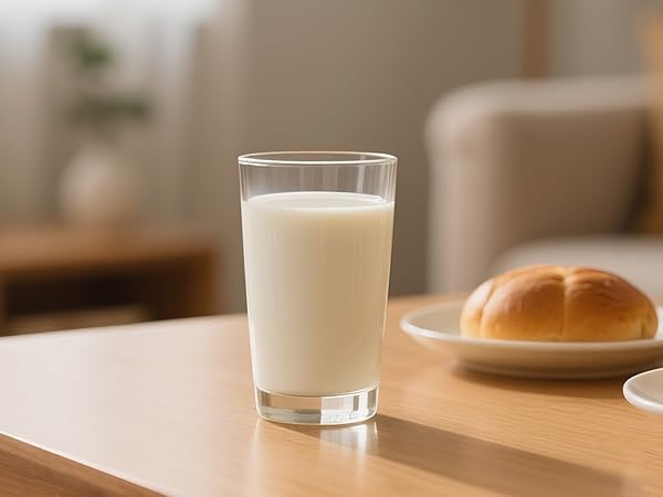 Glass of white or cream-colored liquid on table with bread roll visible on wooden plate in background.