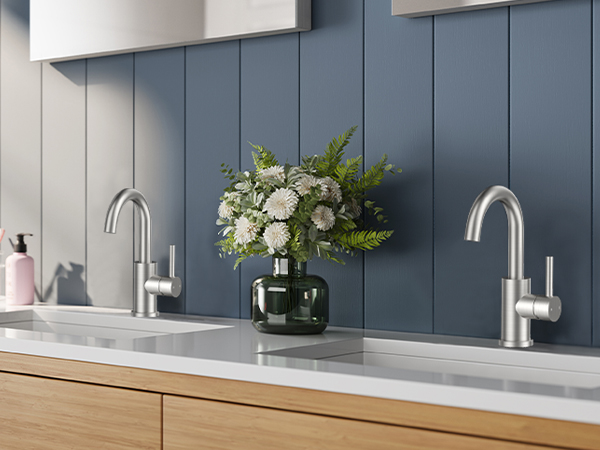 Modern bathroom vanity with dual stainless steel faucets, gray paneled backsplash, and white counter featuring a black vase with fresh flowers.