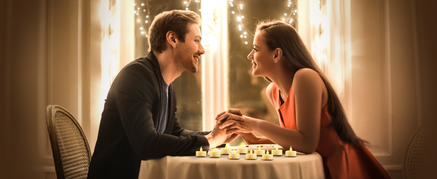 Couple seated at table with chess board, smiling at each other. String lights in background create romantic atmosphere. Man in dark shirt, woman in orange dress.
