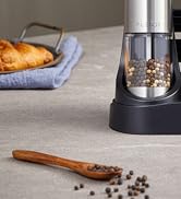 Electric spice grinder with transparent container filled with peppercorns. Wooden spoon with spices nearby, croissant on blue cloth in background.