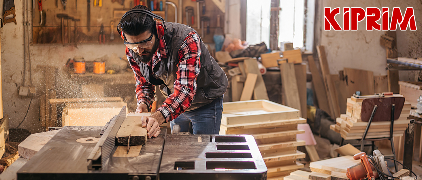 Carpenter working on a wooden project in a workshop, wearing plaid shirt and safety glasses. Various woodworking tools visible in background.
