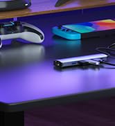 Gaming accessories on a dark surface with purple lighting. A white game controller, blue and pink rectangular objects, and a small device are visible.