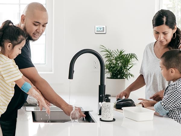 Two parents and two children washing the dishes with a thermostat on the wall behind them.