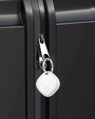 Close-up of a small white circular tracking device attached to a zipper pull on a black luggage or bag. The device has a metallic keyring attachment.