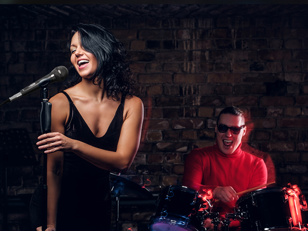 Performance scene in dimly lit venue with musicians on stage against brick wall background, featuring drum kit illuminated with red lighting.