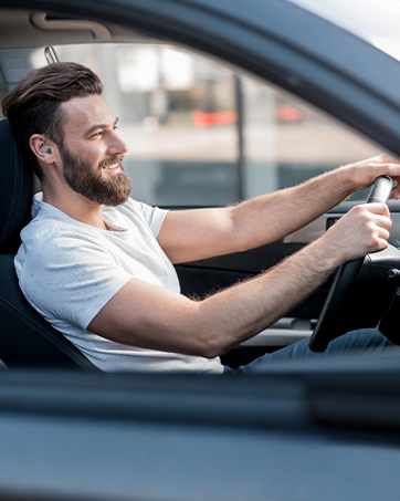 Man with beard wearing light blue shirt driving car, hands on steering wheel, focused expression, daytime outdoor setting with blurred background.