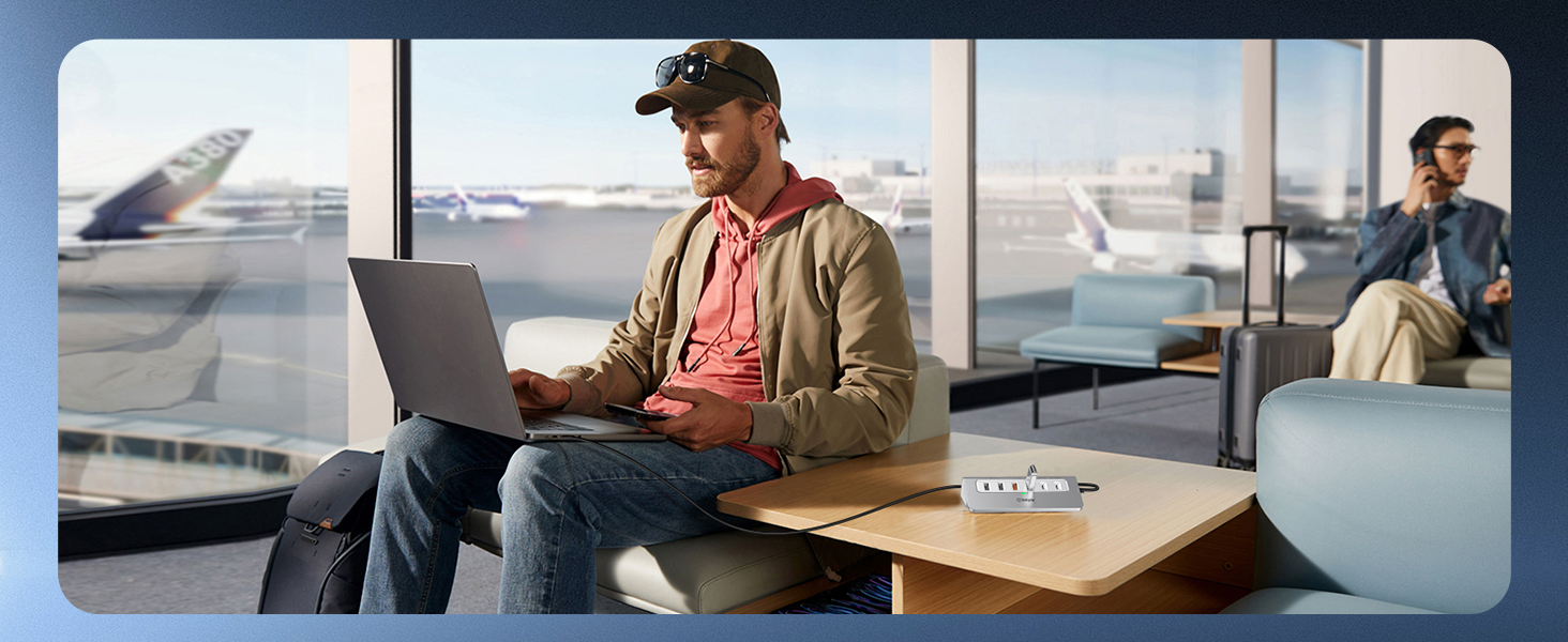 Man using laptop in airport lounge. Casually dressed in cap, jacket, and jeans. Airplanes visible through large windows. Another person in background using phone.